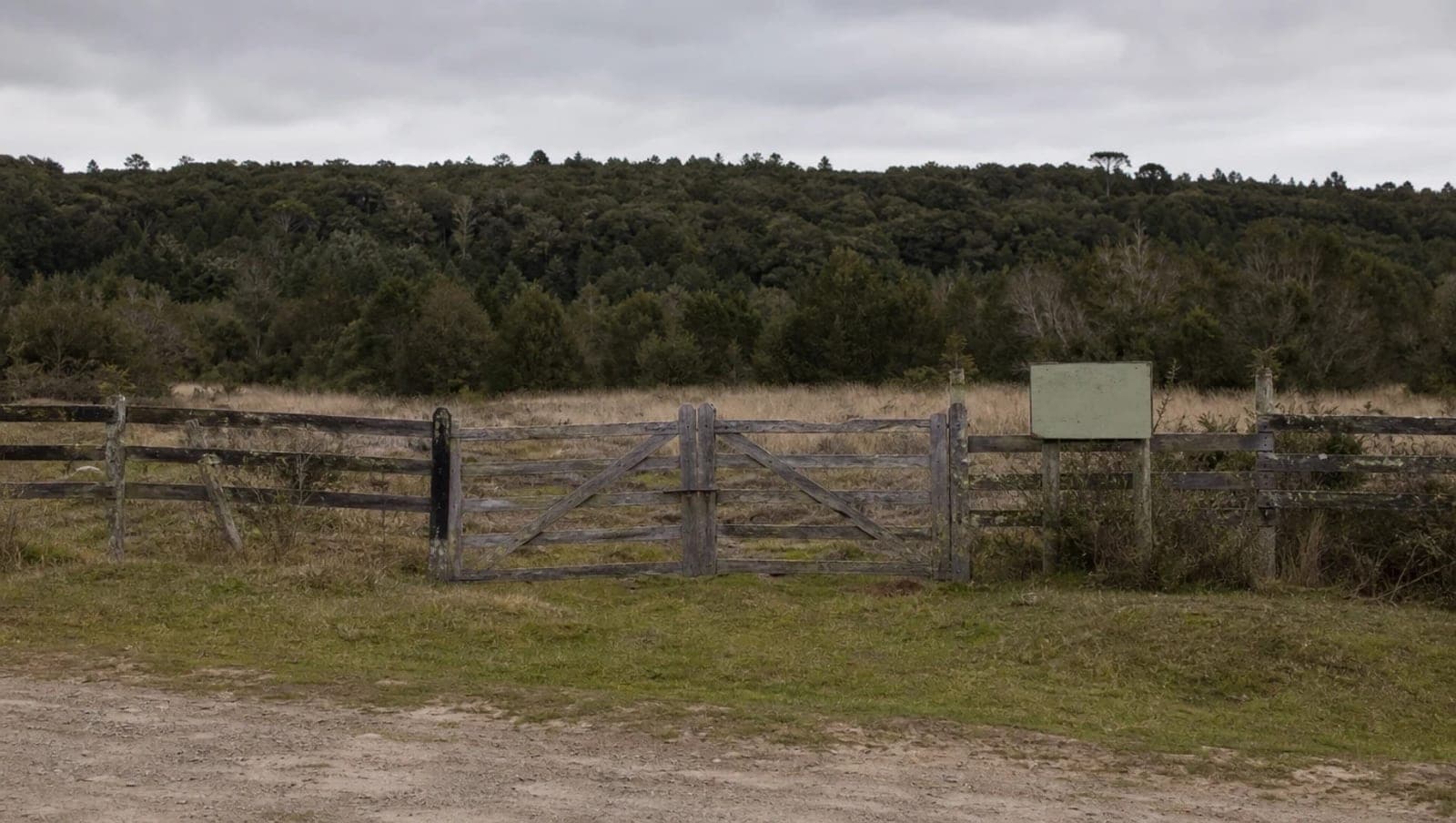 Terreno rural vacío con cerco de madera, portón cerrado y un cartel de venta descolorido, con bosque nativo al fondo